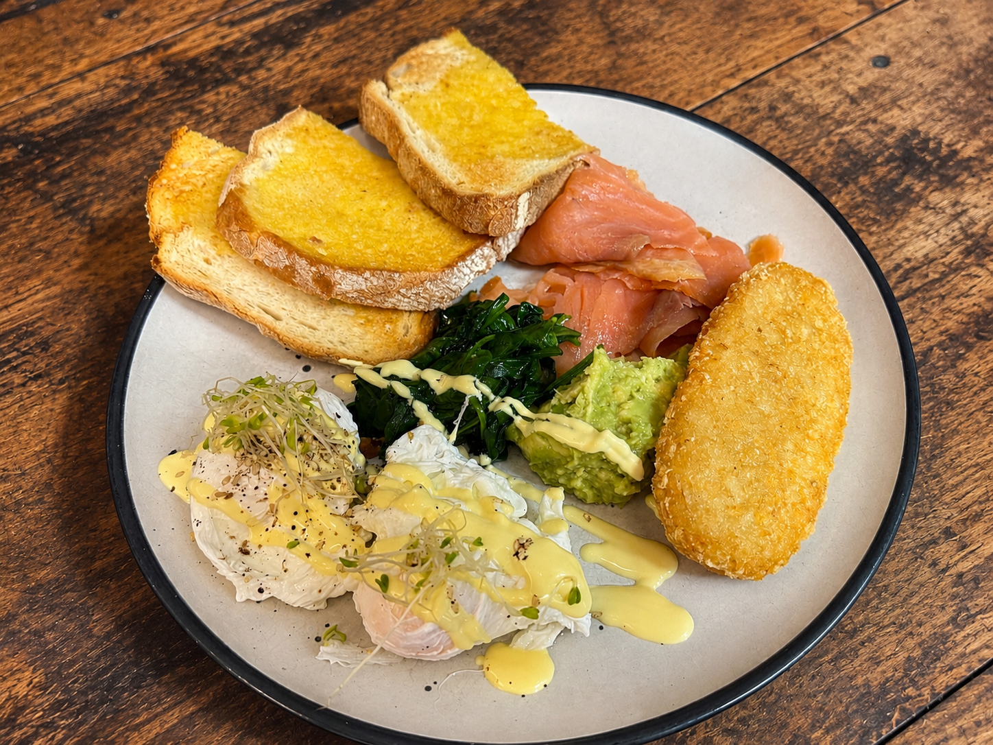 Full breakfast plate with smoked salmon, avocado, hash brown, and poached egg on rustic timber table