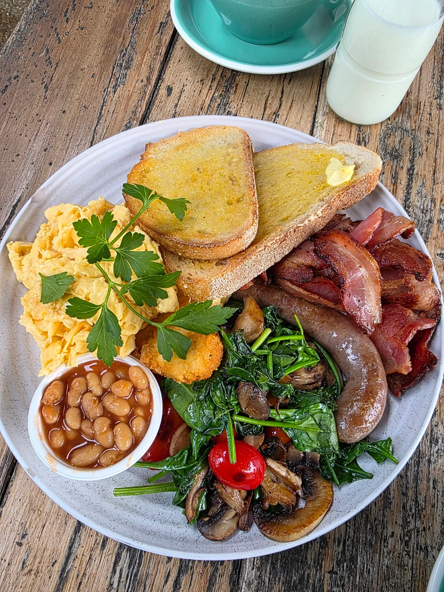 Generous full cooked breakfast with bacon, eggs, toast, sausage and greens on white plate